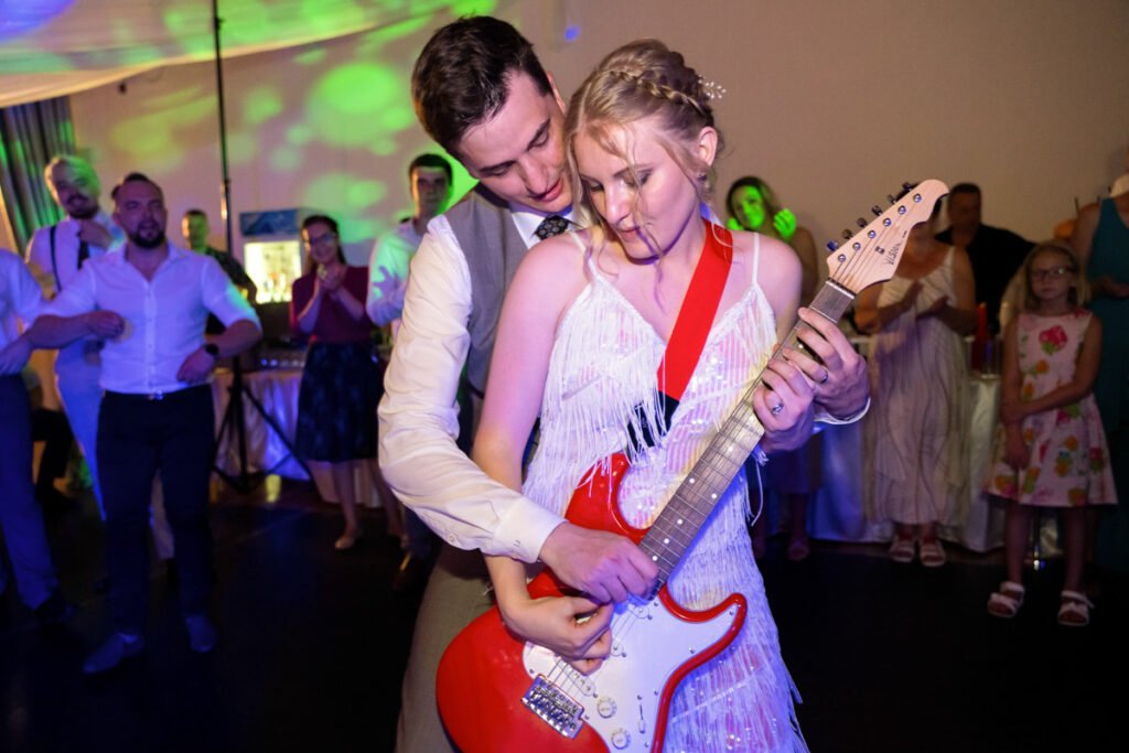 Esküvői ceremóniafotós által megörökített különleges pillanat egy hajdúnánási lakodalmon. A képen az ifjú pár egy piros elektromos gitáron játszik, miközben a vendégek figyelik őket. A menyasszony bohém rojtos ruhában és vörös gitárpánttal tartja a hangszert, míg a vőlegény gyengéden segíti a játékot. A háttérben színes fények és ünneplő vendégek fokozzák a bulihangulatot. Az esküvőfotós kreatív látásmódja kiemeli a rock’n’roll tematikát és az egyedi hangulatot.