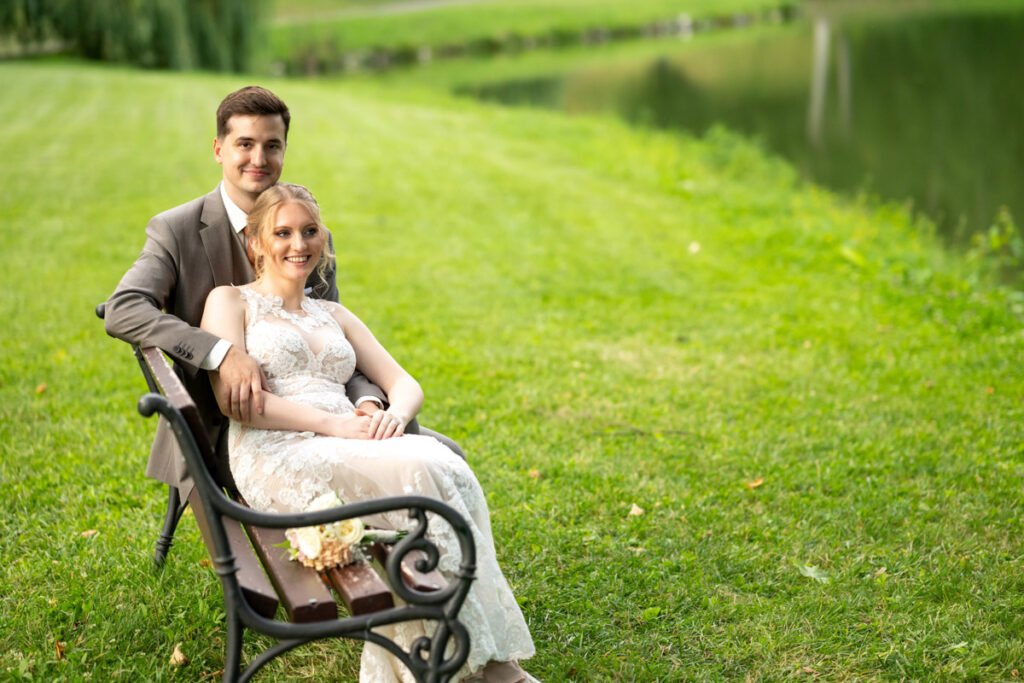 Egy boldog ifjú pár ül egy kovácsoltvas paddon a füzérradványi Károlyi-kastély tóparti kertjében. A menyasszony csipkés, elegáns esküvői ruhában pihen a vőlegény karjaiban, miközben kezében egy finom virágcsokor nyugszik. A vőlegény szürke öltönyben, mosolyogva öleli menyasszonyát, a háttérben pedig a természet zöldje és a tó tükröződő vízfelülete látható. Ez a meghitt és természetközeli esküvői fotó tökéletes példája egy profi esküvői fényképész által készített kreatív jegyesfotózásnak, amely a szerelmesek közti érzelmeket és a varázslatos helyszín szépségét örökíti meg.