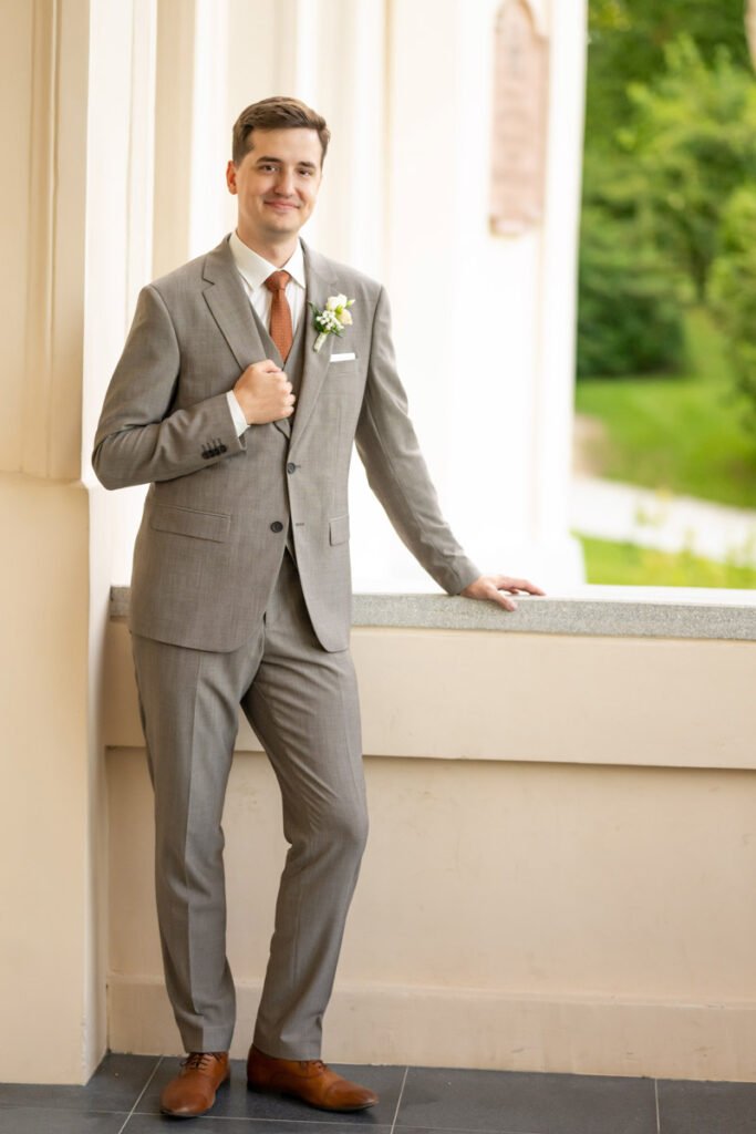 Egy elegáns vőlegény áll a füzérradványi Károlyi-kastély árkádos folyosóján, elegáns szürke öltönyben és barna bőrcipőben. Mellkasán finom virágdísz ékesíti zakóját, nyakkendője pedig harmonizál az esküvői stílusával. Magabiztosan és mosolyogva pózol, egyik kezével zakóját fogva, míg a másikkal könnyedén támaszkodik a kőkorlátra. A természetes fény kiemeli az elegáns megjelenést, és egy időtálló esküvői portrét eredményez. Egy profi esküvői fotográfus által készített stílusos és karakteres vőlegényfotó, amely tökéletes kiegészítője az esküvői kreatív fotózásnak.