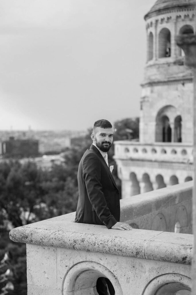 A vőlegény klasszikus fekete-fehér esküvői portréja a Halászbástya elegáns erkélyén Budapesten. Az öltönyös férfi határozottan tekint a kamerába, miközben a háttérben Budapest ikonikus látképe sejlik fel. Ez a prémium esküvői fényképezés tökéletesen visszaadja az eleganciát és a nagy nap időtlen hangulatát.