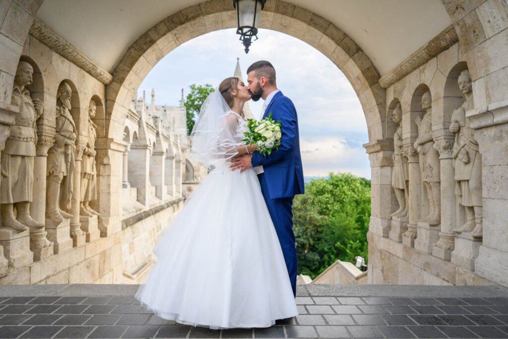 Esküvői fotós - Egy szerelmes ifjú pár csókot vált a Halászbástya egyik impozáns kőboltíve alatt, ahol a kreatív esküvői fotózás Budapest történelmi szépségével fonódik össze. A menyasszony klasszikus fehér esküvői ruhát és fátylat visel, kezében elegáns virágcsokorral, míg a vőlegény stílusos kék öltönyben öleli őt. A háttérben a Halászbástya részletgazdag szobrai és Budapest panorámája teszik különlegessé ezt a romantikus esküvői fényképezést.