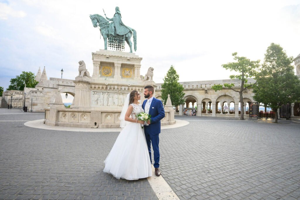 Egy ifjú pár áll a Halászbástya központi terén, Szent István király szobra előtt, ahol a profi esküvői fotográfus Budapest egyik legszebb helyszínén örökíti meg szerelmüket. A menyasszony lenyűgöző fehér csipkés esküvői ruhát és fátylat visel, kezében elegáns virágcsokorral. A vőlegény klasszikus kék öltönyben áll mellette, gyengéden megérintve kezét. A háttérben a történelmi Halászbástya boltívei és a város lenyűgöző panorámája látható, tökéletes környezetet biztosítva egy kreatív esküvői fényképész számára. Ez az exkluzív esküvői portréfotózás a Budai Vár egyik legikonikusabb pontján készül, ahol a ceremóniafotós művészi látásmódja időtálló emléket teremt.