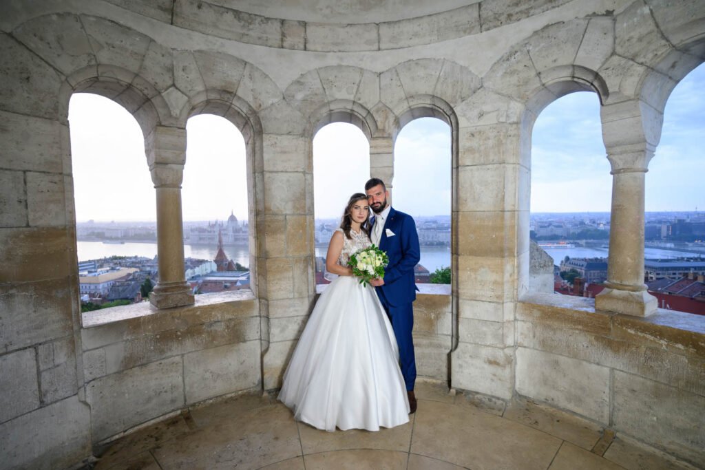 Egy ifjú házaspár áll a Halászbástya kőablakai között, mögöttük a Duna és az Országház lenyűgöző látványa. A menyasszony elegáns fehér ruhájában, kezében virágcsokorral simul vőlegénye mellé, aki stílusos kék öltönyt visel. A kreatív esküvői fotózás Budapesten különleges fényképekkel örökíti meg a nagy nap romantikus pillanatait.