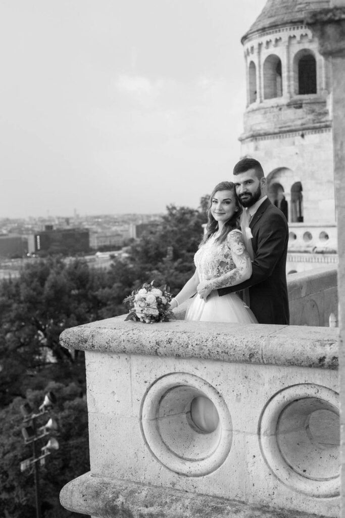 A vőlegény gyengéden átöleli a menyasszonyt a Halászbástya egyik korlátjánál, miközben Budapest lenyűgöző panorámája látható a háttérben. A fekete-fehér esküvői fényképezés időtlen hangulatot kölcsönöz a pillanatnak, kiemelve a pár eleganciáját és az érzelmek mélységét. A kreatív esküvői portréfotózás tökéletes választás a romantikus budapesti esküvői helyszínek szerelmeseinek.