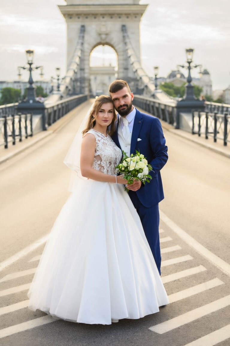 Eskővi fotós - Egy friss házaspár áll Budapest ikonikus hídjának közepén, elegánsan és boldogan. A menyasszony klasszikus fehér esküvői ruhát visel csipkés felsőrésszel és fátyollal, kezében fehér rózsákból készült csokrot tart. A vőlegény sötétkék öltönyben, fehér ingben és nyakkendőben öleli őt.