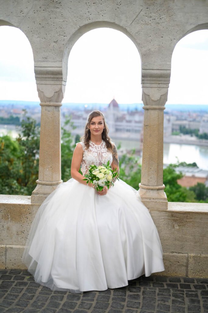 Egy gyönyörű menyasszony hófehér esküvői ruhában ül a Halászbástya elegáns kőkorlátján, kezében friss virágcsokorral. A háttérben Budapest ikonikus látképe, a Parlament és a Duna teszi felejthetetlenné ezt a prémium esküvői fényképezést. Az exkluzív esküvői fotózás tökéletesen megörökíti a menyasszony eleganciáját és a nagy nap romantikus hangulatát.