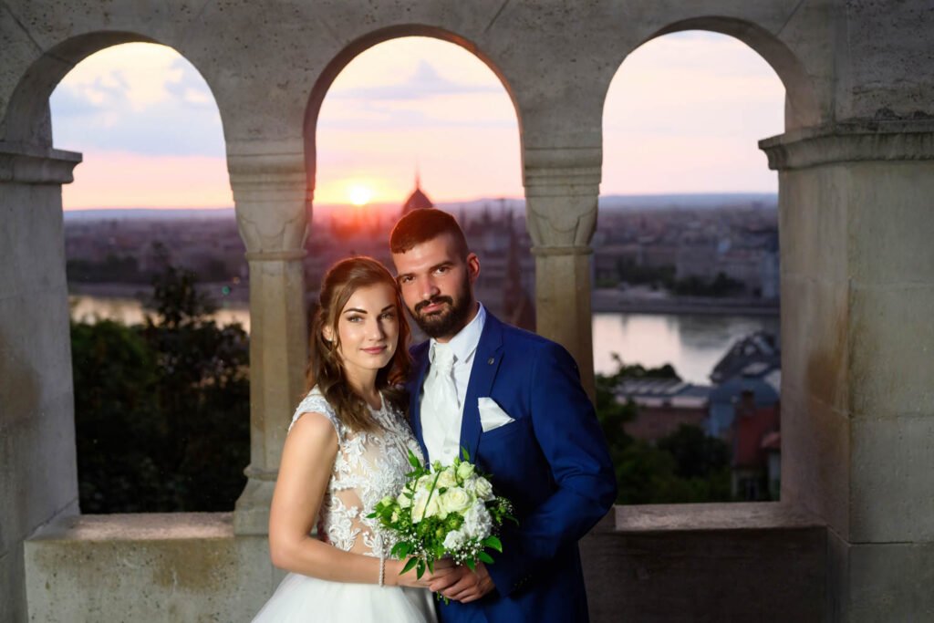 Egy friss házaspár áll a Halászbástya ikonikus oszlopai között, miközben a naplemente aranyfénye bevilágítja Budapest panorámáját. A menyasszony hófehér esküvői ruhájában és a vőlegény elegáns kék öltönyében romantikus pillanatot osztanak meg. A kreatív esküvői fényképezés Budapesten tökéletes helyszínt talál a Halászbástya történelmi hangulatában, ahol a páros fotózás minden részlete emlékezetessé válik.