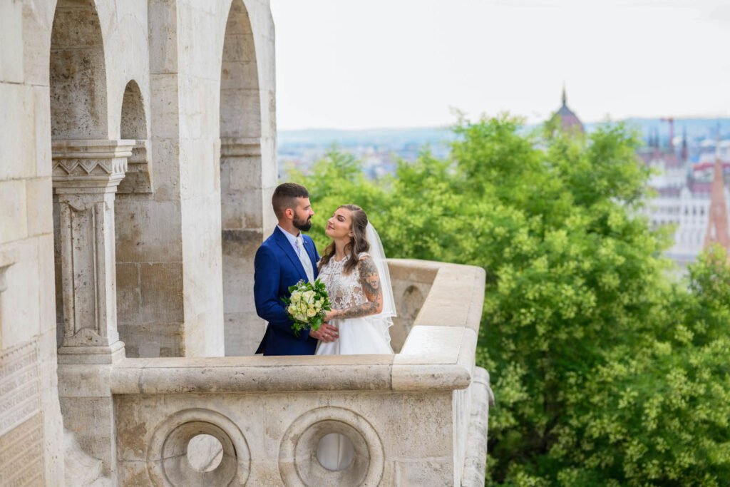 Egy szerelmes ifjú pár áll a Halászbástya egyik elegáns erkélyén, ahonnan lenyűgöző kilátás nyílik Budapest ikonikus épületeire és a Duna-parti látképre. A menyasszony csipkés fehér esküvői ruhát és fátylat visel, kezében gyönyörű fehér rózsacsokrot tart, miközben szerelmesen néz a vőlegényére. A vőlegény elegáns kék öltönyben és fehér ingben áll mellette, meghitt pillanatot osztva meg vele. A Halászbástya mesebeli kőoszlopai és boltívei tökéletes helyszínt biztosítanak egy időtlen esküvői fényképsorozathoz. Exkluzív esküvőfotózás Budapesten, elegáns páros portrék és felejthetetlen emlékek profi esküvői fényképésszel.
