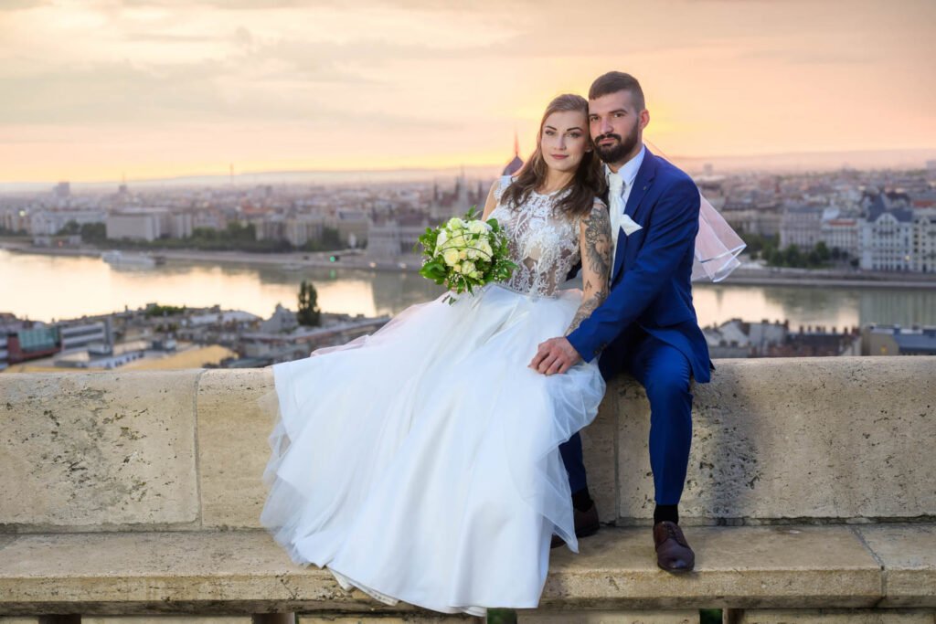 A menyasszony és a vőlegény a Halászbástya egyik kőkorlátján ülve élvezik a naplemente varázslatos fényét Budapest felett. A menyasszony elegáns csipkés esküvői ruhában, kezében fehér rózsacsokorral mosolyog a kamerába, míg a vőlegény kék öltönyben öleli őt. A háttérben a Duna és a Parlament épülete teszi teljessé a romantikus esküvői fényképezés exkluzív hangulatát.