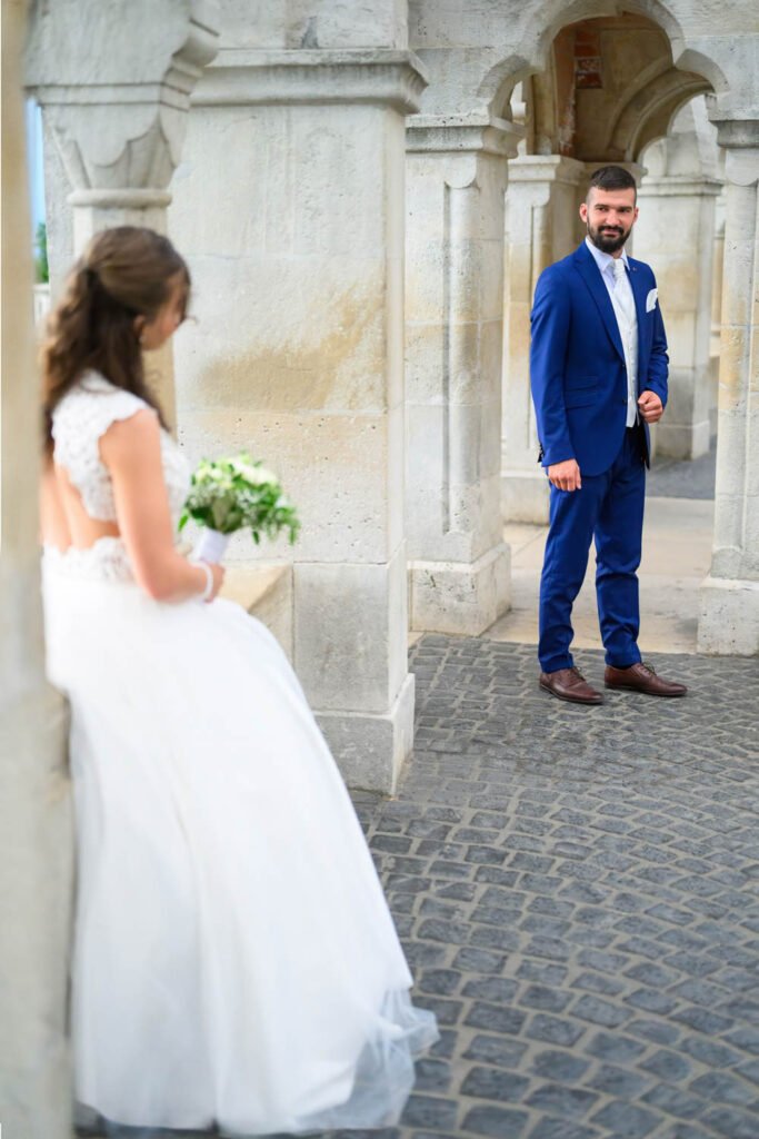 A menyasszony és a vőlegény romantikus pillanatot osztanak meg a Halászbástya elegáns boltívei alatt Budapesten. A menyasszony fehér csipkeruhában virágcsokrot tart, miközben a vőlegény kék öltönyben mosolyogva tekint rá. Ez a kreatív esküvői fényképezés Magyarország egyik legszebb esküvői fotós helyszínén örökíti meg a szerelem időtálló pillanatait.