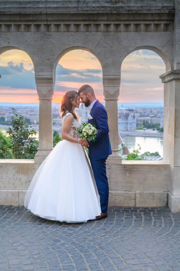 A menyasszony és a vőlegény Budapest egyik legszebb esküvői fotós helyszínén, a Halászbástya boltívei alatt állnak, miközben a naplemente varázslatos fénye megvilágítja őket. A kreatív esküvői fényképezés során a pár gyengéd pillanatot oszt meg, miközben a háttérben a Parlament épülete és a Duna csodás panorámája látható. Ez a prémium esküvői portréfotózás tökéletesen megörökíti a szerelem időtlen szépségét.