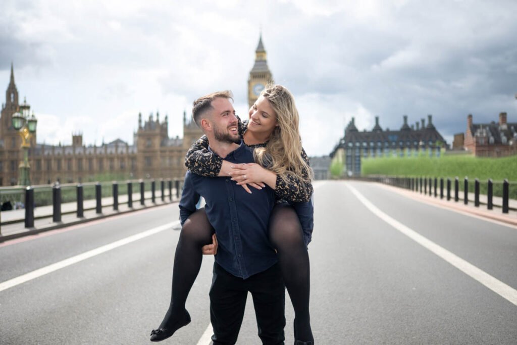 Romantikus jegyesfotózás Londonban – Egy boldog szerelmespár vidám pillanata a Westminster hídon, a Big Ben ikonikus hátterével. Kreatív és természetes esküvői fotózás eljegyzéshez és pároknak.
