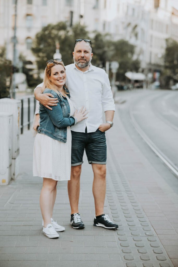 Esküvői fényképész Budapesten – Egy boldog pár ölelkezik Budapest utcáin, háttérben villamossínekkel és városi épületekkel. A jegyesek lezser, de elegáns öltözetben állnak a kamera előtt: a menyasszony fehér ruhát és farmerdzsekit visel, míg a vőlegény fehér inget és farmer rövidnadrágot. Az esküvői fotográfus célja, hogy a jegyespár természetes pillanatait és a városi hangulatot is megörökítse.