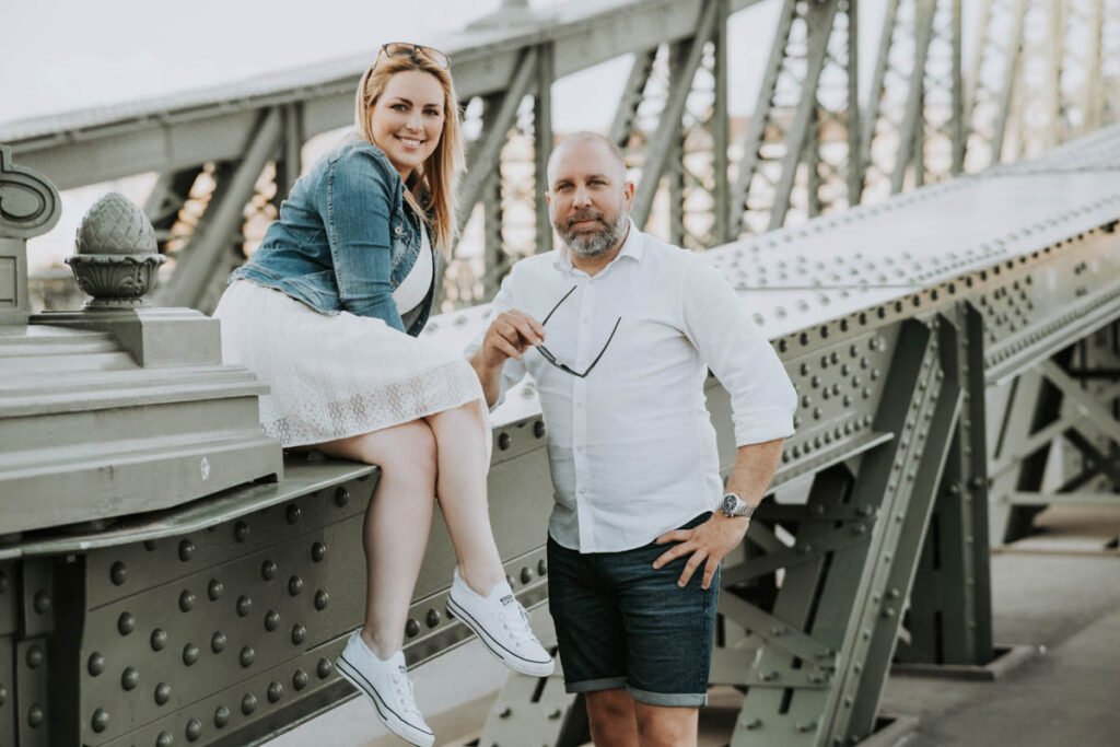 Budapesti esküvői fotós által készített jegyesfotó a Szabadság híd ikonikus fém szerkezetén. A menyasszony fehér ruhában és farmerdzsekiben, sportos cipőben ül a híd korlátján, miközben boldogan mosolyog. A vőlegény mellette áll elegáns fehér ingben és sötét rövidnadrágban, kezében napszemüveggel. A város panorámája és a különleges helyszín tökéletes hátteret nyújt egy romantikus esküvői kreatív fotózáshoz Budapesten.