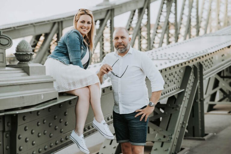 Budapesti esküvői fotós által készített jegyesfotó a Szabadság híd ikonikus fém szerkezetén. A menyasszony fehér ruhában és farmerdzsekiben, sportos cipőben ül a híd korlátján, miközben boldogan mosolyog. A vőlegény mellette áll elegáns fehér ingben és sötét rövidnadrágban, kezében napszemüveggel. A város panorámája és a különleges helyszín tökéletes hátteret nyújt egy romantikus esküvői kreatív fotózáshoz Budapesten.
