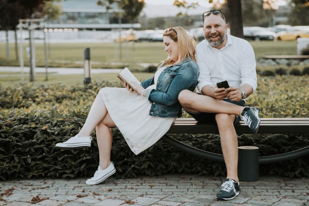 Esküvői blog - Egy szerelmes pár egy parkban egy modern padon ülve nevetgél. A nő fehér ruhában és farmerdzsekiben könyvet olvas, miközben mosolyogva támaszkodik párjára. A férfi fehér inget és farmer rövidnadrágot visel, egyik lábát keresztbe téve a padon, miközben a telefonját tartja és vidáman nevet. A háttérben autók és városi környezet látható, mégis meghitt és természetes pillanatot örökít meg ez a kreatív páros fotózás. Profi esküvői fotográfus, jegyesfotózás, eljegyzési portrék és romantikus esküvő előtti fényképezés élettel teli pillanatokkal.