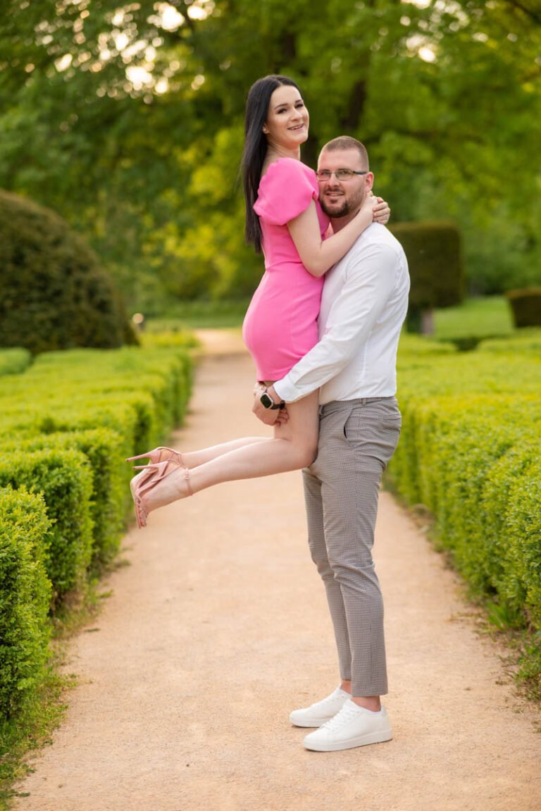 Esküvői fotózás Nyíregyházán – Egy szerelmes pár örömteli pillanata egy gyönyörű, gondozott kertben. A vőlegény elegáns fehér inget és szürke nadrágot visel, miközben mosolyogva tartja karjaiban menyasszonyát, aki egy feltűnő rózsaszín ruhában ragyog. A menyasszony kecsesen átkarolja kedvesét, miközben boldogan néz a kamerába. A buja zöld bokrok és fák között kanyargó sétaút tökéletes helyszínt biztosít az esküvő előtti romantikus fotók elkészítéséhez.