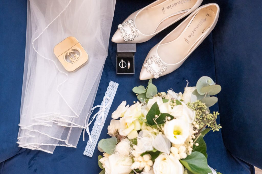 Művészi flatlay kompozíció menyasszonyi kellékekről királykék bársony háttéren, krémszínű hegyes orrú balerina cipő kristály csattal díszítve, fehér tüll fátyol szegéllyel, arany Gucci parfüm doboz, gyémánt eljegyzési gyűrű fekete bársony dobozban, fehér csipke harisnyakötő, fehér menyasszonyi csokor kálákból és rózsákból eukaliptusz levelekkel és apró fehér virágokkal díszítve, esküvői részletfotós Budapest, kreatív kellékfotós, luxus esküvői fotóművész, prémium részletfotó specialista, vintage stílusú esküvői fotóriporter, elegáns pillanatok megörökítője, menyasszonyi készülődés fotográfus