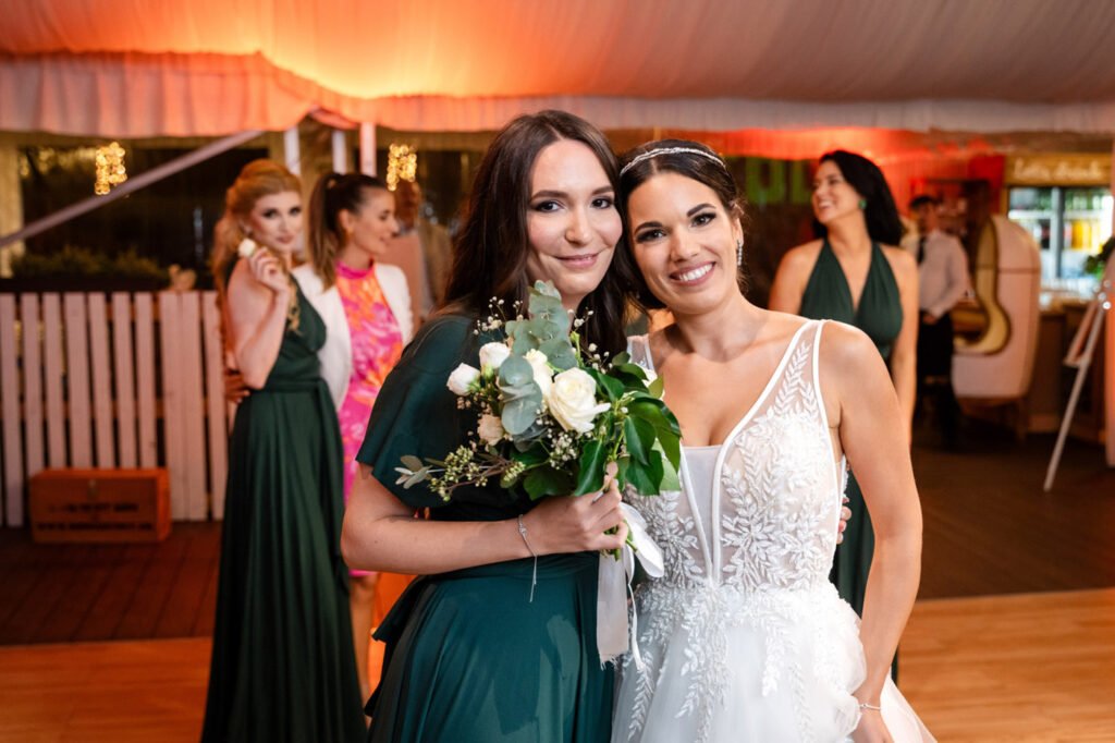 Örömteli esküvői pillanatkép a Wedding Wood rendezvényhelyszínen, a menyasszony és a szerencsés csokorkapó közös fotója, menyasszony levélmintás csipke applikációval díszített, V-kivágású fehér menyasszonyi ruhában, kristályos fejdísszel és konttyal, mellette a csokorkapó smaragdzöld hosszú estélyi ruhában, kezében a fehér rózsás-eukaliptuszos menyasszonyi csokor, háttérben narancssárgás hangulatvilágítás és fehér drapériák, koszorúslányok zöld és rózsaszín ruhákban, fa parkettás táncparkett, esküvői pillanatfotós Budapest, kreatív party fotográfus, művészi esküvői riporter, prémium ceremóniafotós, luxus rendezvényfotós, spontán pillanatok megörökítője