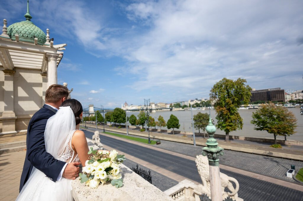 Egy szerelmes ifjú pár romantikus pillanatát örökíti meg a profi esküvői fotós Budapest lenyűgöző panorámájával a háttérben. A vőlegény elegáns sötétkék öltönyben öleli menyasszonyát, aki egy csodás uszályos fehér esküvői ruhát és fátylat visel. Kezük ügyében egy gyönyörű fehér virágcsokor pihen a kőkorláton. A Lánchíd, a Duna és a pesti belváros ikonikus épületei teszik még varázslatosabbá ezt a meghitt pillanatot. Ez a kép tökéletes példája az érzelmeket megörökítő prémium esküvői fényképezésnek, amely egyedi és időtálló emlékeket teremt a nagy napról.