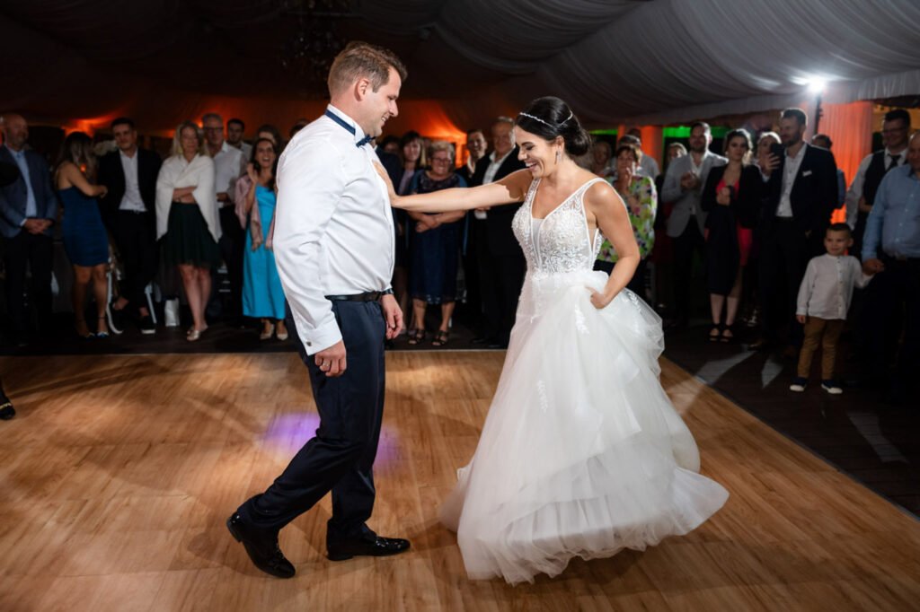 Dinamikus esküvői pillanatkép a Wedding Wood rendezvényhelyszínen, vidám első tánc közben, menyasszony csipke díszítésű, V-kivágású, pántos fehér menyasszonyi ruhában tüll szoknyarésszel, kristályos fejdísszel és kontyba tűzött frizurával, vőlegény fehér ingben, csokornyakkendőben és sötétkék nadrágban, fa parkettás táncparketten táncolnak, háttérben vendégek körben állva, türkizkék, sötétkék ruhákban, narancssárgás dekorvilágítás és fehér drapériák a sátorban, esküvői party fotós Budapest, művészi pillanatfotós, kreatív ceremóniafotós, első tánc specialista, luxus rendezvényfotográfus, prémium esküvői történetmesélő, spontán pillanatok megörökítője