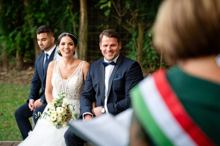 Szabadtéri szertartás - Menyasszony és vőlegény boldogan mosolyog az esküvői szertartáson a Wedding Wood erdei helyszínén, miközben az anyakönyvvezető beszédet mond. Természetközeli esküvői fotózás varázslatos hangulattal.