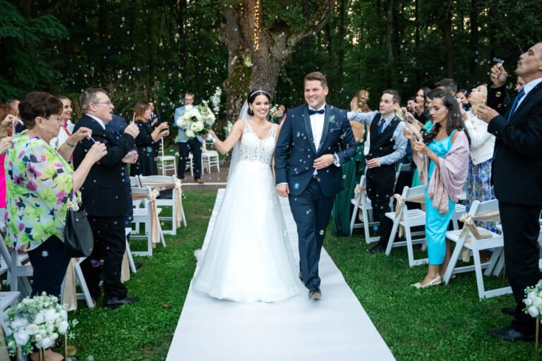 A menyasszony és a vőlegény mosolyogva vonul ki az esküvői szertartás után a Wedding Wood természetközeli helyszínén, miközben a vendégek buborékokat fújnak és ünneplik őket.