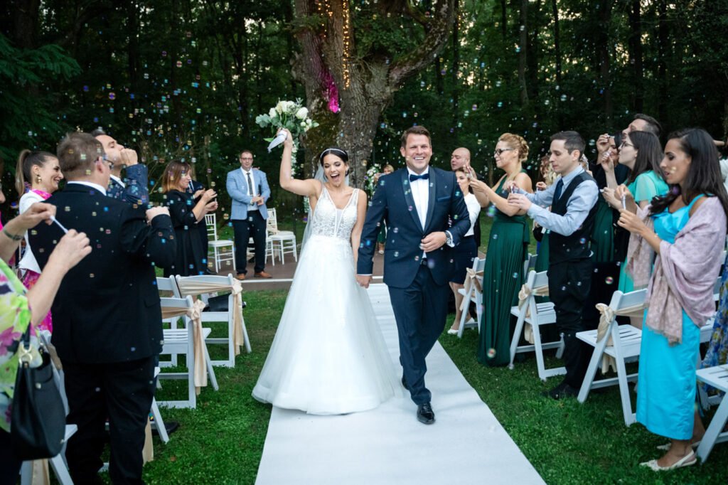 Menyasszony és vőlegény örömmel vonul ki az esküvői szertartás után a Wedding Wood helyszínén, miközben a vendégek buborékokkal ünneplik őket.