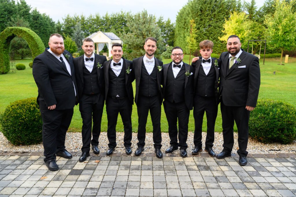 A vőlegény tanúi elegáns öltönyben, összekarolva és nevetve állnak össze egy közös fotóra, megörökítve barátságuk és az esküvői nap különleges hangulatát.
