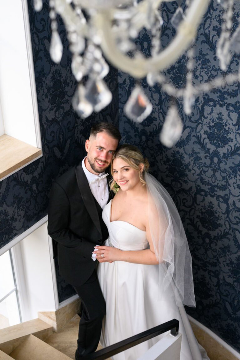 Esküvői történetek -Az ifjú pár elegánsan pózol a Le Til Kúria belső lépcsőházában. A menyasszony hófehér ruhájában és a vőlegény klasszikus fekete öltönyében mosolyognak a kamera felé, miközben a csillár kristálydíszei sejtelmesen keretezik a pillanatot.