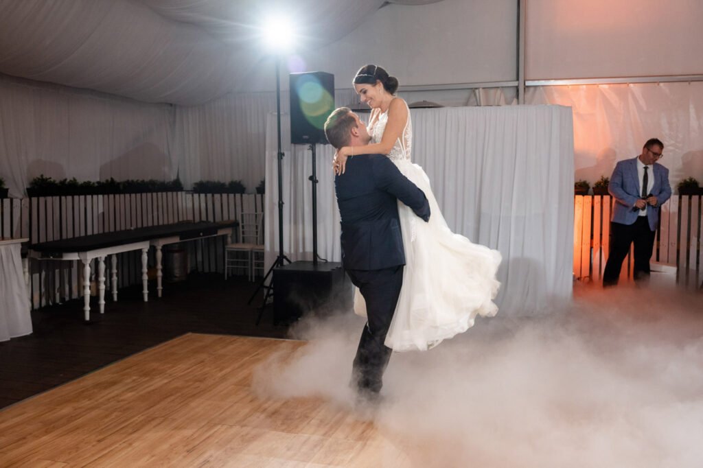 Látványos első tánc pillanatképe a Wedding Wood rendezvényhelyszínen, vőlegény magasba emeli menyasszonyát, menyasszony mély V-kivágású, csipke felsőrészű, tüll szoknyás menyasszonyi ruhában, kontyba tűzött frizurával, vőlegény sötétkék slim fit öltönyben, a párt körülölelő fehér szárazjég-köd látványos fényhatással, háttérben fehér drapériák és narancssárgás dekorvilágítás, fa parkettás táncparkett, háttérben világoskék öltönyös CM, fehér vintage stílusú bárasztalok, esküvői party fotós Budapest, művészi pillanatfotós, kreatív ceremóniafotós, első tánc specialista, luxus rendezvényfotográfus, prémium esküvői történetmesélő, hangulatos pillanatok megörökítője