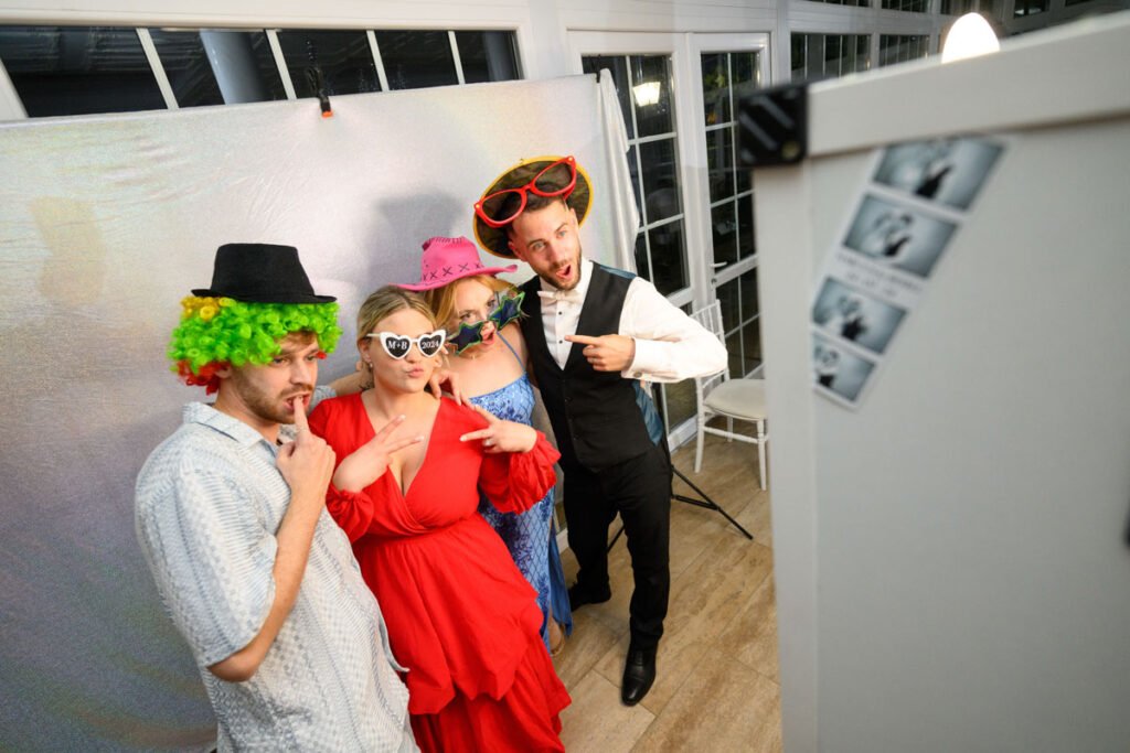 Vidám vendégek színes jelmezekben és vicces kiegészítőkkel pózolnak a selfiebox előtt egy esküvői bulin. A háttérben csillogó dekoráció és a selfiebox által készített fotócsíkok teszik a pillanatot emlékezetessé.