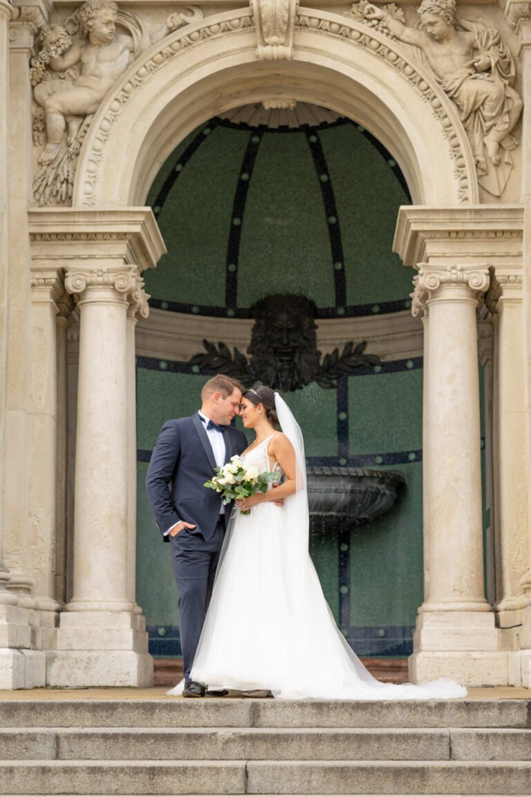 Egy boldog ifjú pár romantikus pillanatát örökíti meg a kreatív esküvői fényképész Budapesten. A vőlegény elegáns sötétkék öltönyt visel csokornyakkendővel, míg a menyasszony gyönyörű uszályos fehér ruhában, fátyollal és virágcsokorral áll mellette. A háttérben egy impozáns, klasszikus stílusú épület díszes oszlopokkal, szobrokkal és egy történelmi szökőkúttal teszi varázslatossá ezt a meghitt esküvői portrét. A fotó tökéletes példája az exkluzív esküvői fényképezésnek, amely a természetes érzelmeket és a nagy nap szépségét hangsúlyozza.