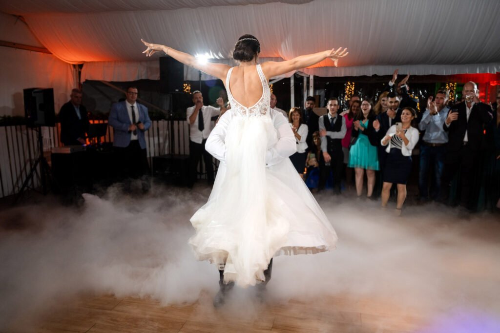 Művészi esküvői pillanatkép a Wedding Wood rendezvényhelyszínen, menyasszony kitárt karokkal táncol a szárazjég-ködben, csipke díszítésű, mély hátú, pántos fehér menyasszonyi ruhában, tüll szoknyarésszel, fején kristályos fejdísz és kontyba tűzött frizura, a háttérben vendégek tapsolnak és ünnepelnek, világoskék öltönyös és nyakkendős úr, fehér inges-mellényes vendég, türkizkék ruhás hölgy vendég látható, narancssárgás dekorvilágítás és fehér drapériák a sátorban, fa parkettás táncparkett, esküvői party fotós Budapest, művészi pillanatfotós, kreatív ceremóniafotós, első tánc specialista, luxus rendezvényfotográfus, prémium esküvői történetmesélő, hangulatos pillanatok megörökítője