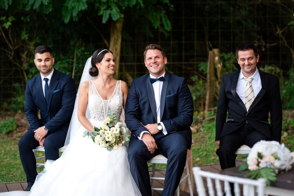 Boldog menyasszony és vőlegény elegáns esküvői öltözetben, mosolyogva a szertartás után. Esküvői fotózás természetközeli helyszínen.