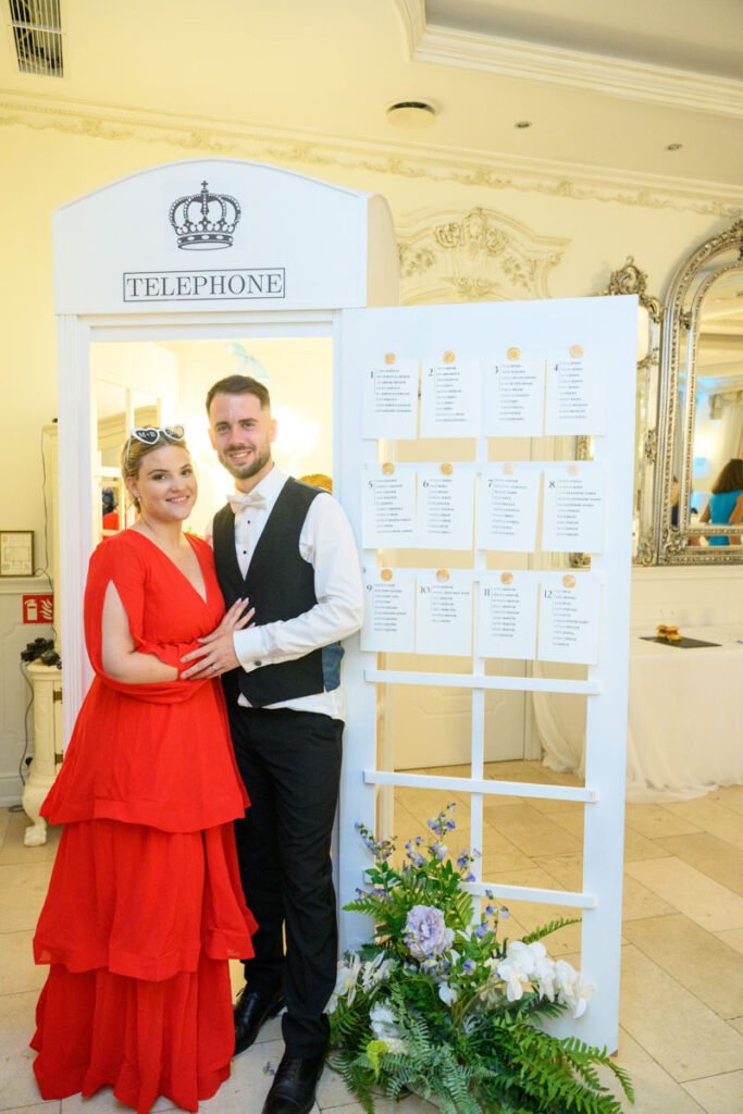 A menyasszony piros ruhában és a vőlegény elegáns mellényben mosolyognak a kamera felé egy esküvői ülésrendet bemutató installáció előtt. A dekoráció virágokkal és részletekkel teszi hangulatossá a jelenetet.
