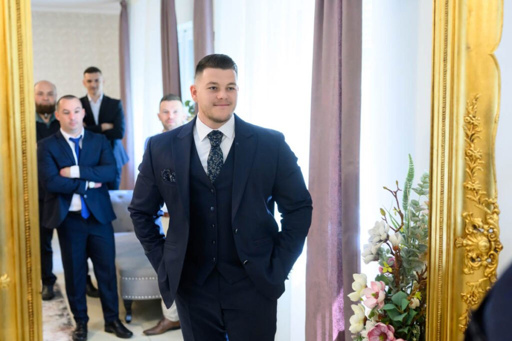 A vőlegény sötétkék háromrészes öltönyben készülődik egy elegáns, aranykeretes tükrökkel díszített teremben, a vőfélyekkel a háttérben