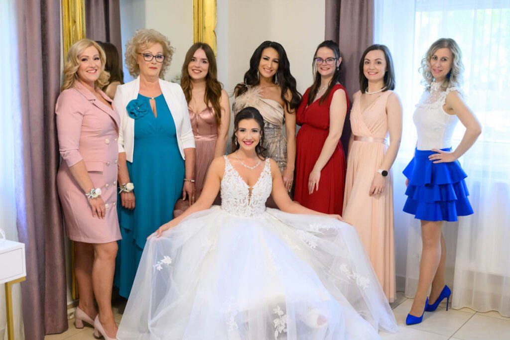 Ülő menyasszony szétterített ruhában, körülötte az édesanyja és a női vendégek elegáns ruhákban a készülődés helyszínén