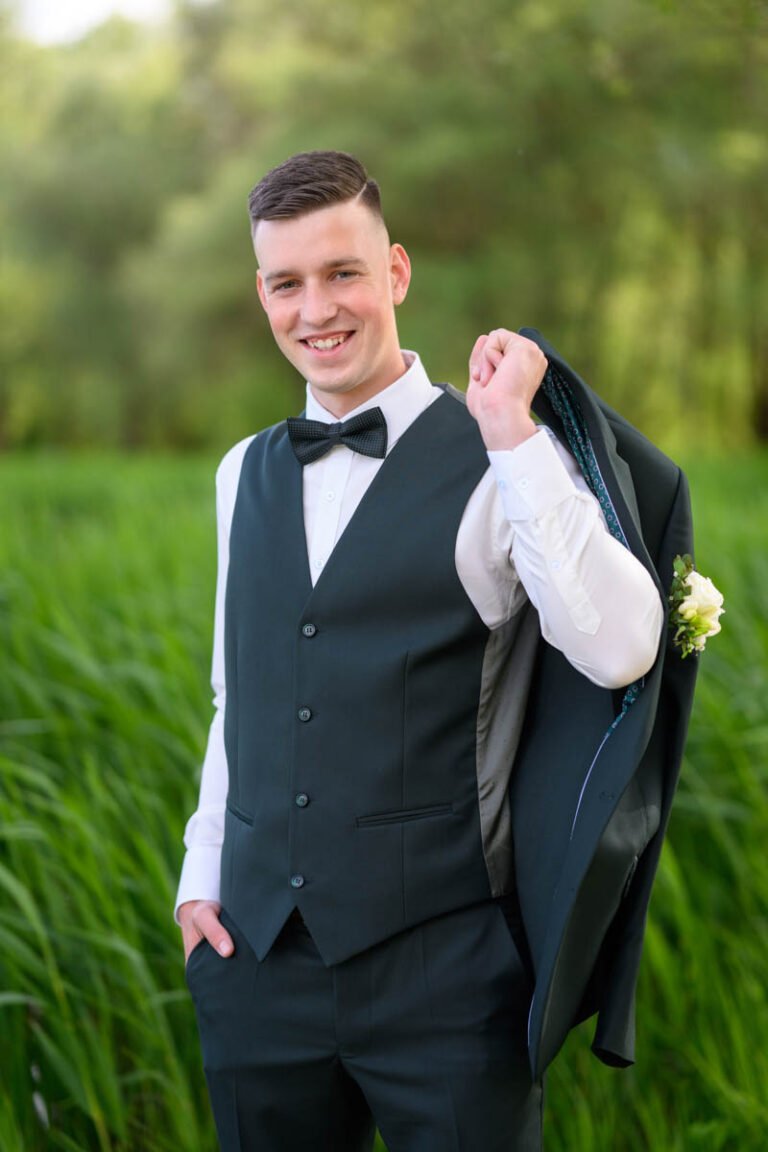 Mosolygó vőlegény mellényben, vállán átvetett zakóval a zöld mező előtt