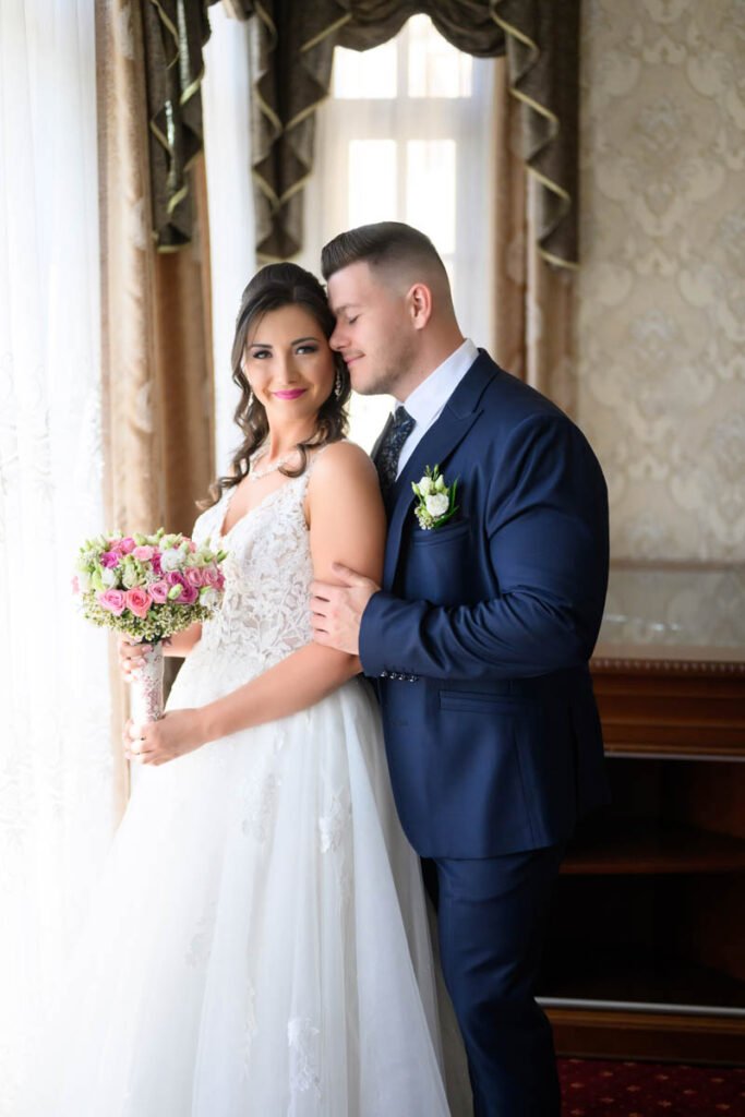 A vőlegény hátulról öleli a menyasszonyt egy elegáns terem ablaka előtt, a menyasszony kezében rózsaszín csokorral