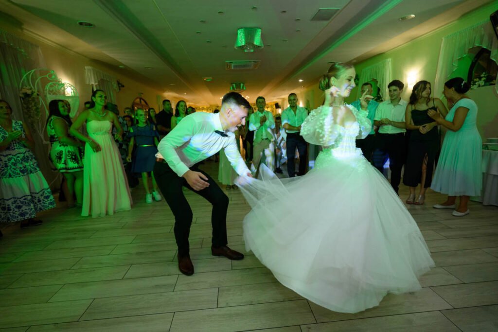 Pörgő menyasszony és táncoló vőlegény a vendégsereg gyűrűjében, esti fényben